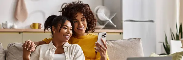 A joyful lesbian couple relaxes on a sofa, sharing laughter while looking at a phone, banner — Stock Photo