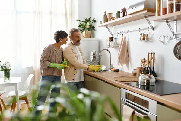 A mature couple enjoys each others company while washing dishes in a sunlit kitchen. — Stock Photo