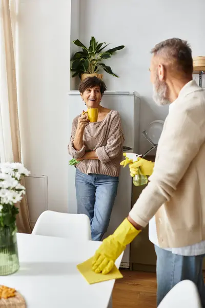 A smiling woman leans against the wall with a cup while a man cleans the table, creating warmth. — Stock Photo