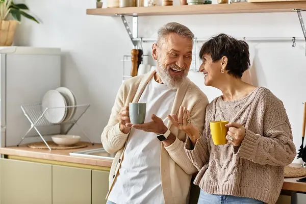A mature couple shares laughs and warm drinks in a bright and inviting kitchen at home. — Stock Photo