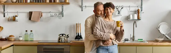 A happy mature couple stands closely in a warm kitchen, savoring a moment over a cup. — Stock Photo