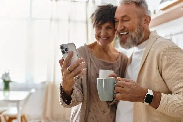 Um casal feliz ri juntos enquanto segurando canecas e olhando para um smartphone em uma cozinha acolhedora. — Fotografia de Stock