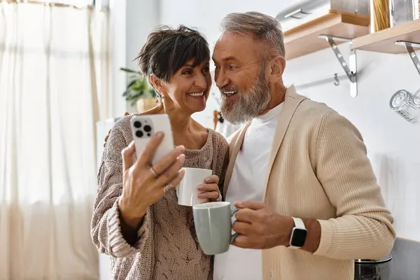Casal sênior compartilha um momento alegre, segurando canecas e capturando memórias em uma cozinha ensolarada. — Fotografia de Stock
