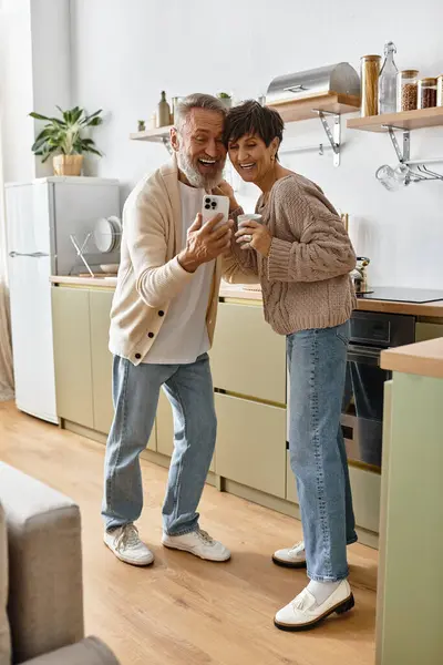 A mature couple shares laughter while taking a selfie in their bright kitchen together. — Stock Photo