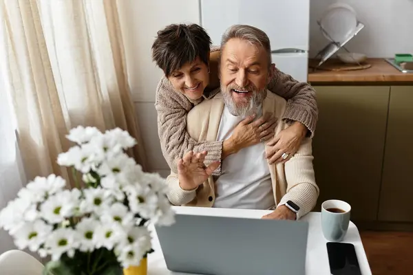 A woman embraces a man from behind as they smile at a laptop in their warm, inviting kitchen. — Stock Photo