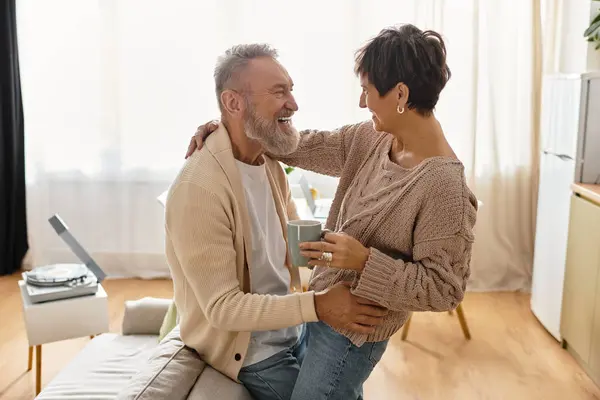 Mature couple embraces and laughs while sipping coffee in their cozy living room — Stock Photo