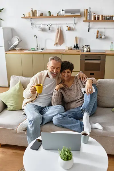 A mature couple laughs together on the sofa while watching something on their laptop. — Stock Photo
