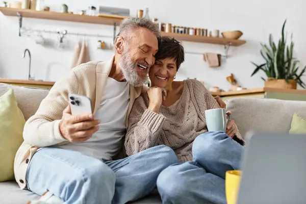 A joyful couple smiles and shares stories while relaxing on their couch with warm beverages. — Stock Photo