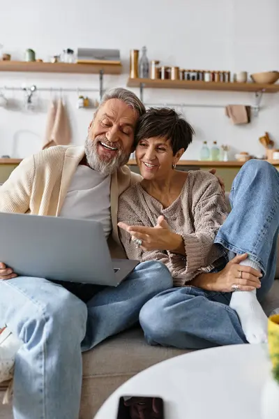 A mature couple sits closely on a couch, excitedly using their laptop in a cozy living room. — Stock Photo