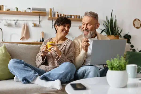 A joyful couple shares laughter and drinks while exploring a laptop in their warm home. — Stock Photo