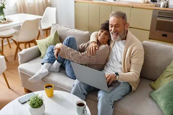 A mature couple sits on a sofa, smiling and sharing joy as they explore content on their laptop. — Stock Photo