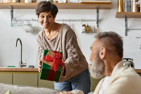 Donna matura presenta un regalo ben avvolto al suo partner sorridente in un accogliente spazio abitativo. — Foto stock