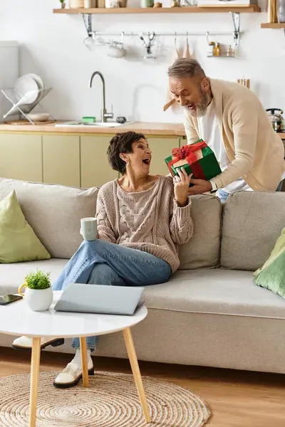 Una coppia felice condivide risate come l'uomo presenta un regalo alla donna nella loro casa elegante. — Foto stock