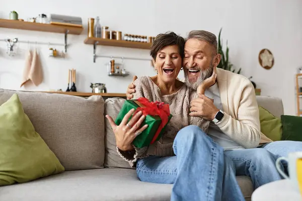 Pareja madura abriendo con entusiasmo un regalo sorpresa mientras se acurruca en su sofá de la sala de estar - foto de stock
