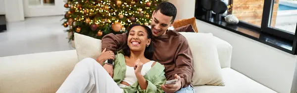 Joyful couple relaxes together on a sofa, surrounded by Christmas decorations and lights, banner — Stock Photo