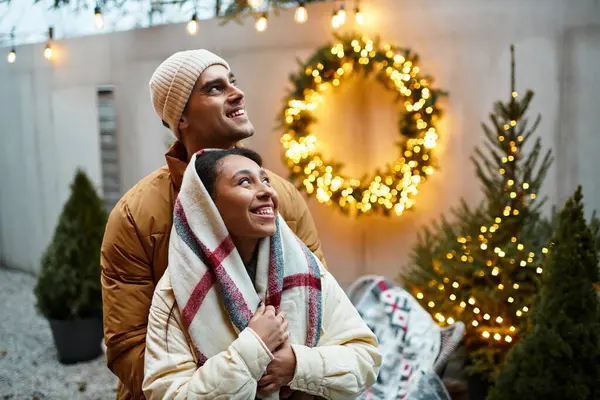 A beautiful couple wraps themselves in warmth, basking in holiday joy surrounded by festive lights. — Stock Photo