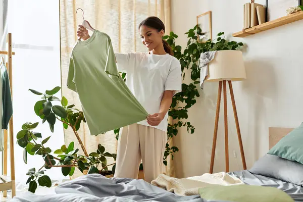 Mulher morena sorri enquanto organiza roupas em um vibrante e moderno espaço de vida cheio de plantas. — Fotografia de Stock