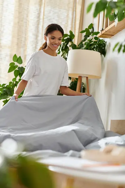 Una joven sonriente hace su cama en un apartamento luminoso y moderno lleno de plantas. - foto de stock