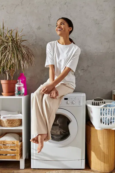 A young African American woman smiles while relaxing on a washing machine in her tidy apartment. — Stock Photo