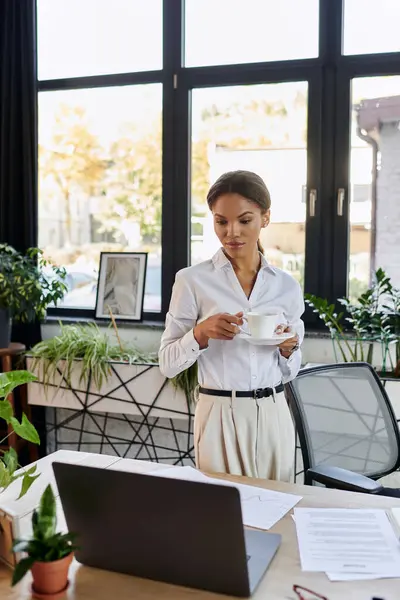 A young african american woman in a white shirt stands by her desk, sipping coffee while working. — Stock Photo