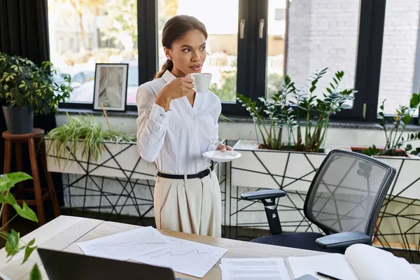 A young African American woman in a white shirt enjoys her coffee while working in an office. — Stock Photo