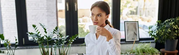 Young African American woman in a white shirt sips coffee while working in a bright office, banner — Stock Photo