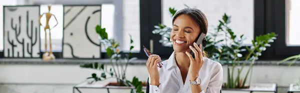 A young Black woman in a white shirt chats animatedly on the phone while working, banner — Stock Photo