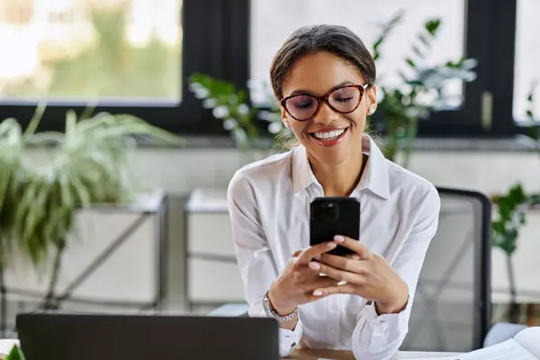 A young African American woman in a white shirt smiles while using her smartphone at work. — Stock Photo