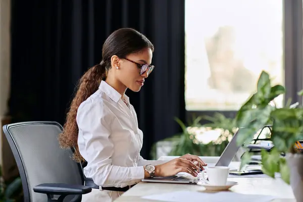 A young African American woman in a white shirt types on a laptop at a sleek, modern office. — Stock Photo