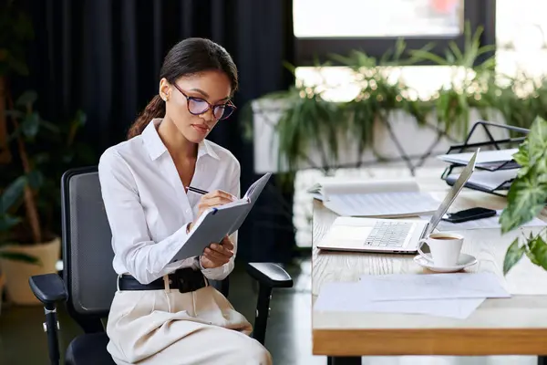 A young african american woman in a white shirt takes notes while seated at a modern office desk. — Stock Photo