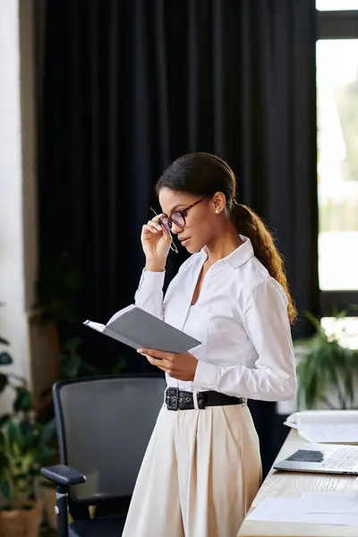 A young African American woman in a white shirt reads intently while standing in an office. — Stock Photo