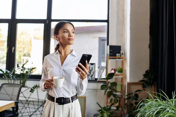 Confident businesswoman holds a cup and smartphone, focused on her work in a modern office space. — Stock Photo