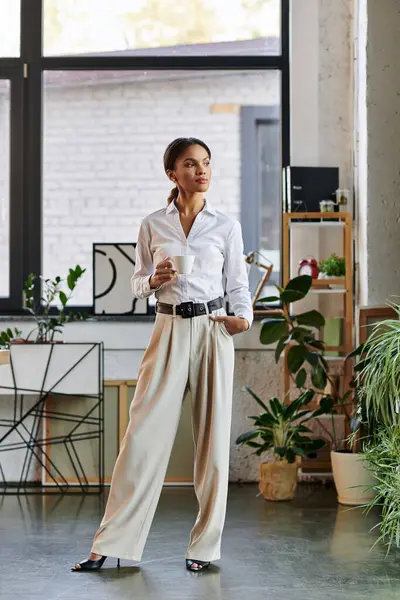 Confident young African American businesswoman stands thoughtfully, holding a cup in bright office. — Stock Photo