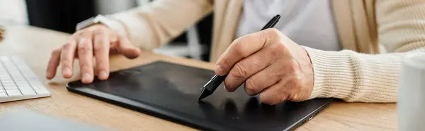 Hombre mayor guapo dibuja en una tableta mientras está sentado en un elegante escritorio de madera. - foto de stock