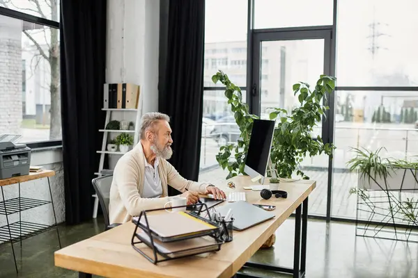Senior man engages in work at a sleek desk surrounded by greenery and natural light. — Stock Photo