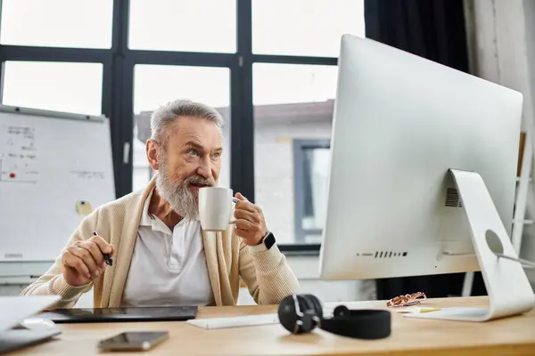 Mature man sips coffee and engages with his computer in a spacious, bright office environment. — Stock Photo