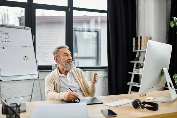 Un uomo bello maturo sta lavorando a una scrivania elegante, tenendo una tazza e scrivendo in un ufficio accogliente. — Foto stock