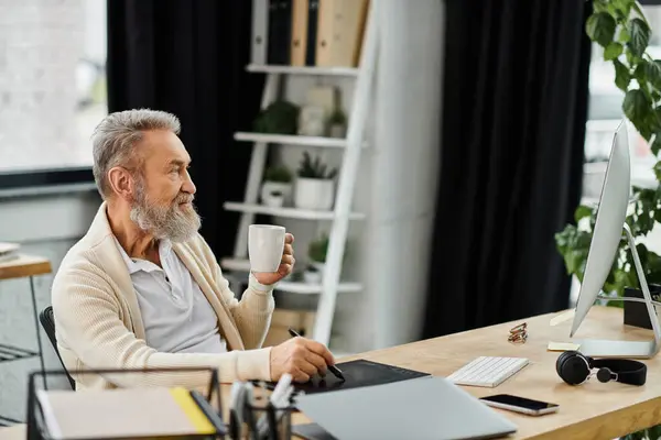 Senior man with a beard sips coffee and focuses on his work in a bright home office setting. — Stock Photo