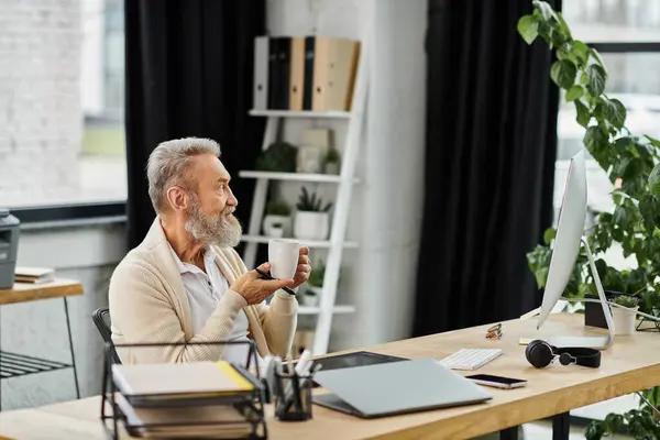 Mature man in a stylish workspace thoughtfully sipping coffee while looking at a computer screen. — Stock Photo