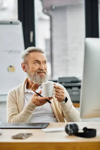 Un uomo bello maturo tiene premurosamente una tazza di caffè mentre è seduto nel suo spazio di lavoro. — Foto stock