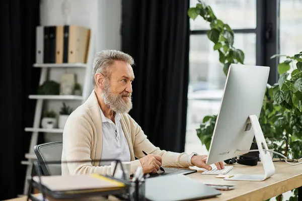 Uomo maturo con la barba che lavora diligentemente al suo computer in un ambiente di ufficio luminoso. — Foto stock