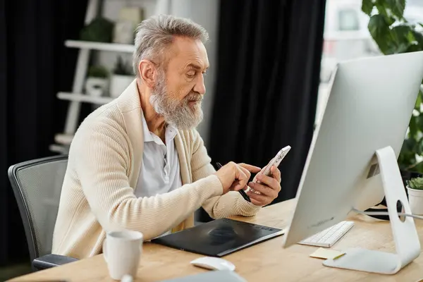 Handsome senior man interacts with his smartphone while seated at a desk with a computer. — Stock Photo