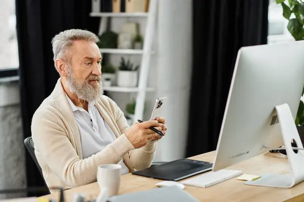 Mature man with beard focuses on smartphone while seated at a desk in a bright office. — Stock Photo