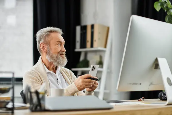 Handsome senior man holds a smartphone, focused on his computer in a modern workspace. — Stock Photo
