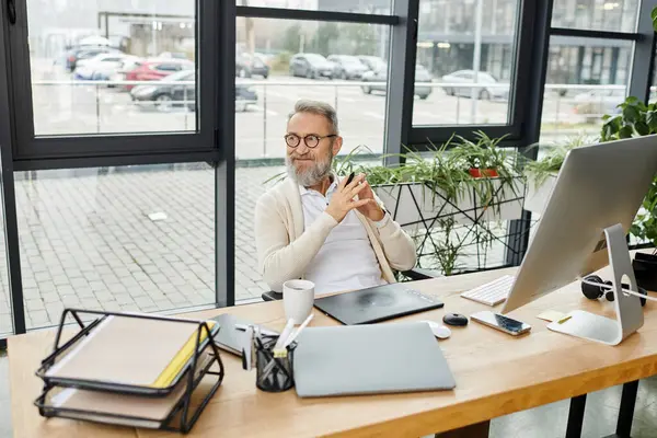 A mature handsome man sits comfortably at a desk, focused on work, surrounded by greenery. — Stock Photo