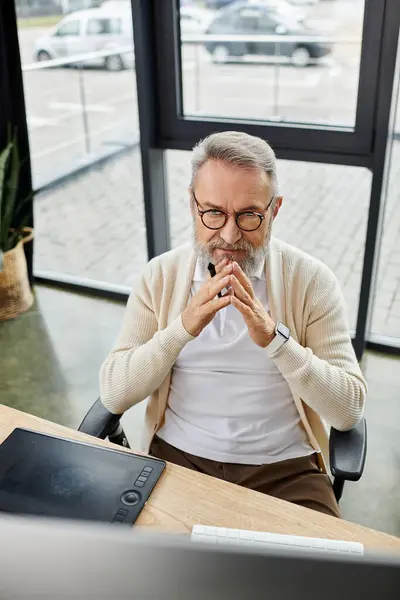 Uomo anziano con occhiali contemplare il lavoro mentre seduto alla scrivania in ufficio contemporaneo. — Foto stock