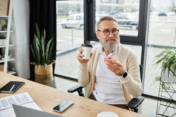 Senior man smiles as he enjoys a cup of coffee and a donut at his desk in a modern workspace. — Stock Photo