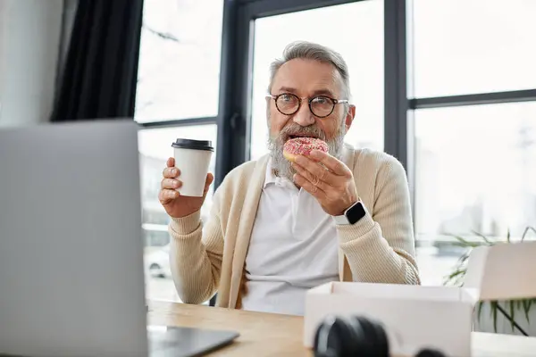 A mature man savoring a donut and coffee at his home workspace near a window. — Stock Photo