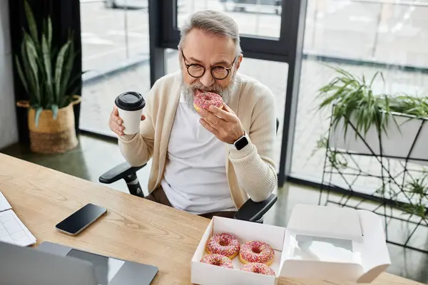 A mature man is savoring a donut while holding coffee at his office desk, surrounded by plants. — Stock Photo