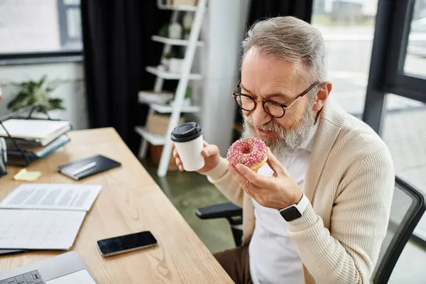 Mature man savoring a donut while holding a cup of coffee in a modern office setting. — Stock Photo